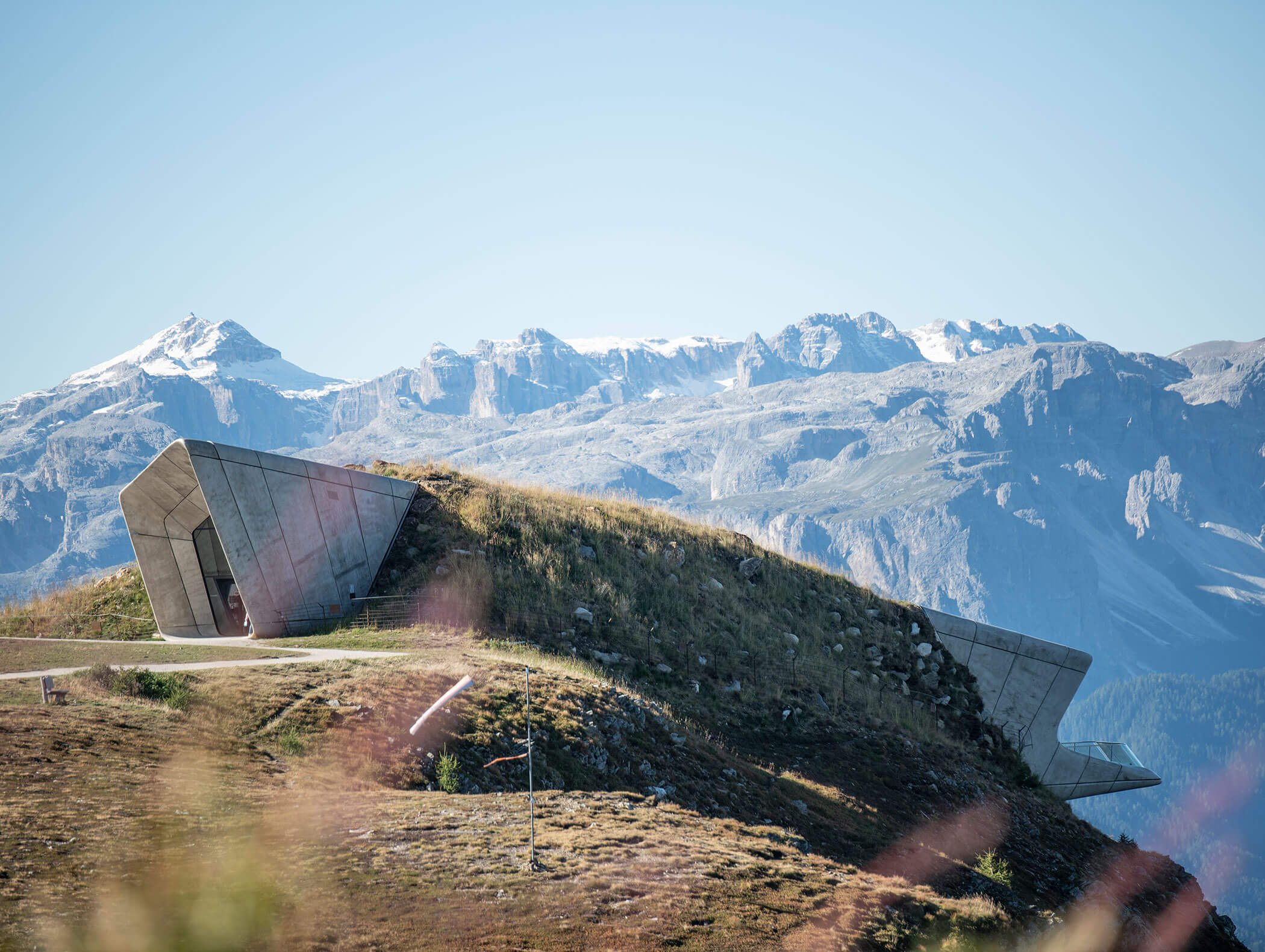 Modernes Betongebäude, das in einen grasbewachsenen Hügel mit Bergen im Hintergrund unter klarem Himmel gebaut wurde. - Majestic Resorts (Relaunch)