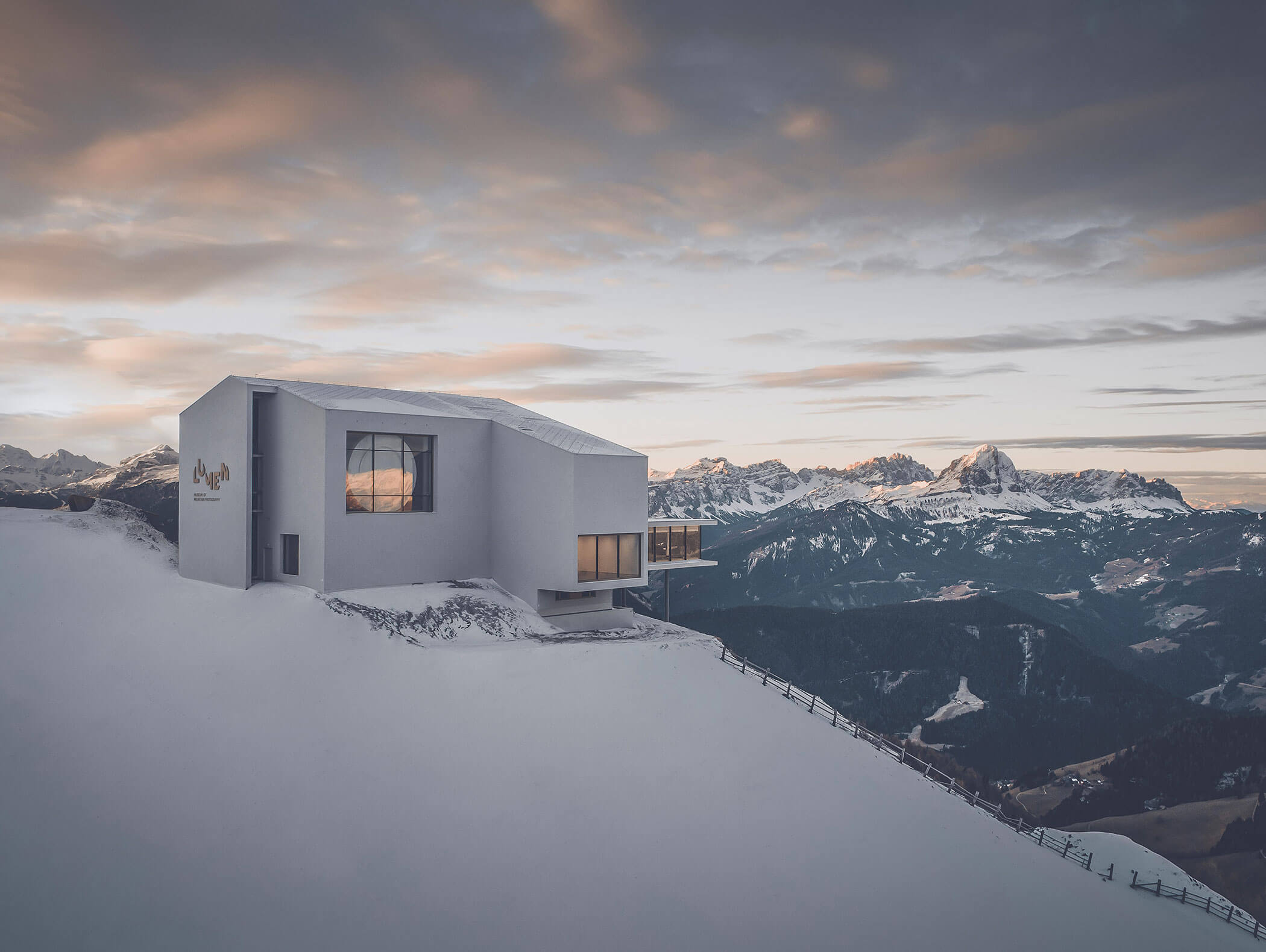 Modernes Gebäude an einem verschneiten Berghang mit entfernten Gipfeln und einem dramatischen Sonnenuntergangshimmel im Hintergrund. - Majestic Resorts (Relaunch)