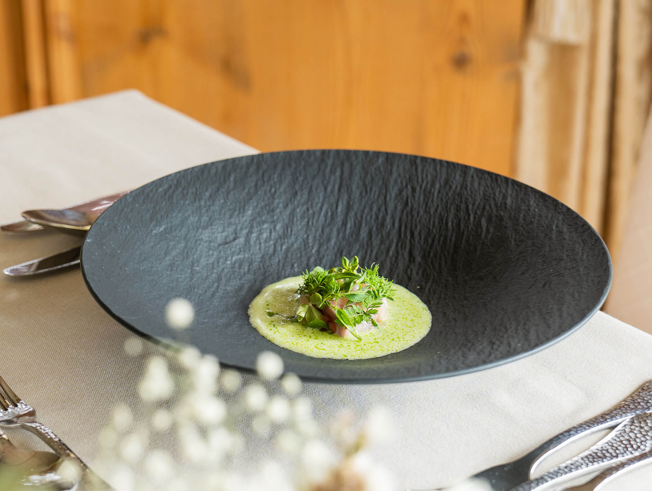 Ein Gourmetgericht mit grüner Soße und Kräutern wird kunstvoll auf einem großen schwarzen Teller auf einem gedeckten Esstisch angerichtet. - Majestic Resorts (Relaunch)