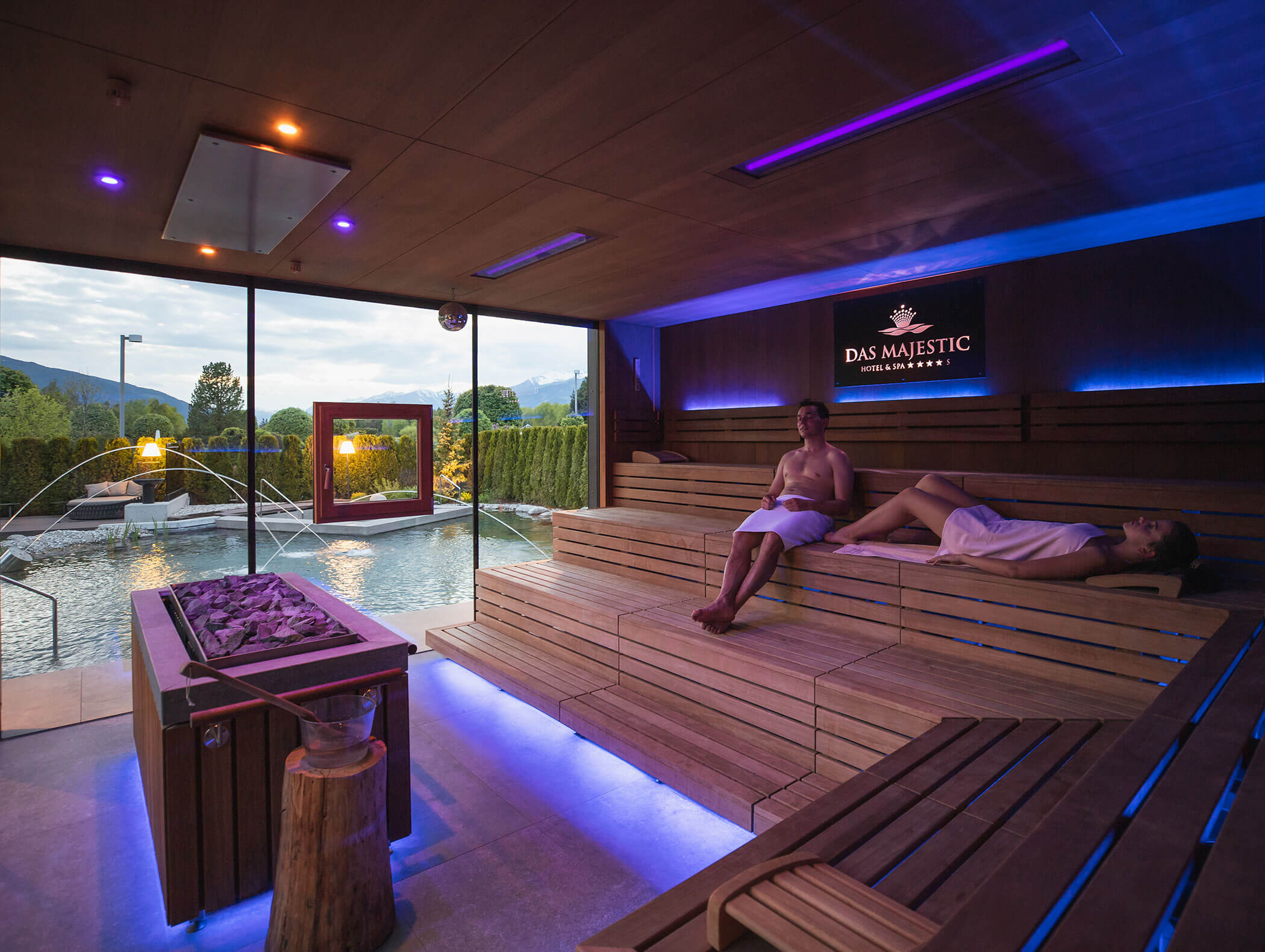 Zwei Personen entspannen in einer modernen Sauna, die durch große Fenster einen Blick auf den Außenpool und die Berge bietet. - Majestic Resorts (Relaunch)