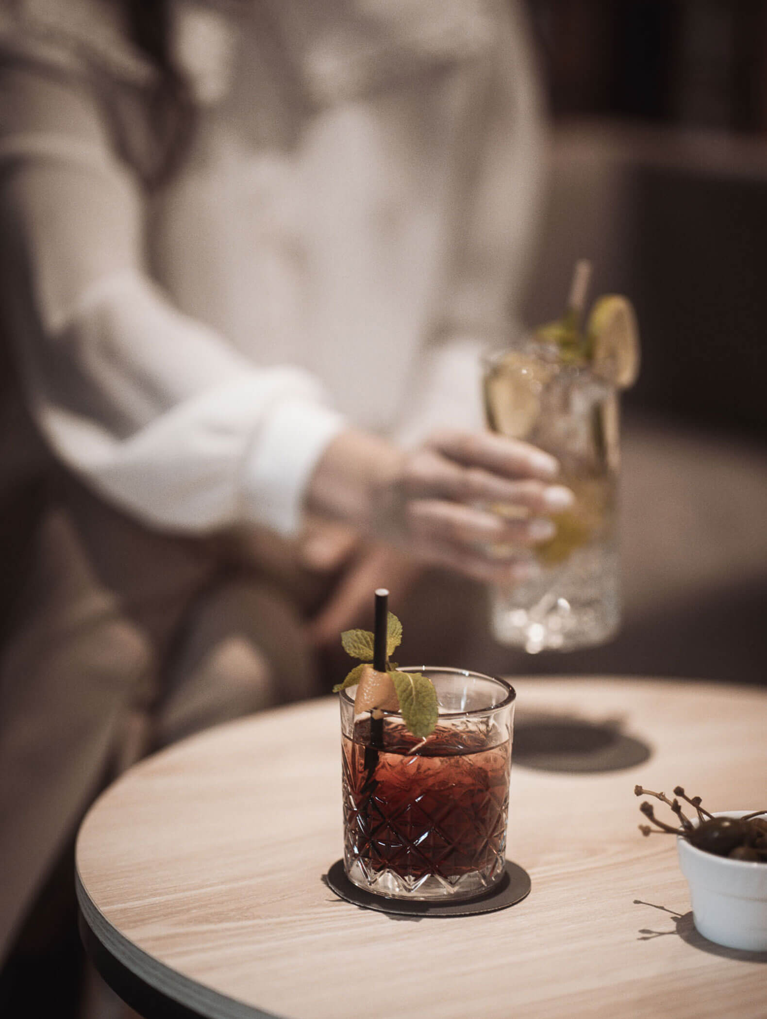 Eine Person nimmt einen Cocktail mit Limette in die Hand, ein anderer Cocktail mit Minze steht auf einem runden Holztisch. - Majestic Resorts (Relaunch)