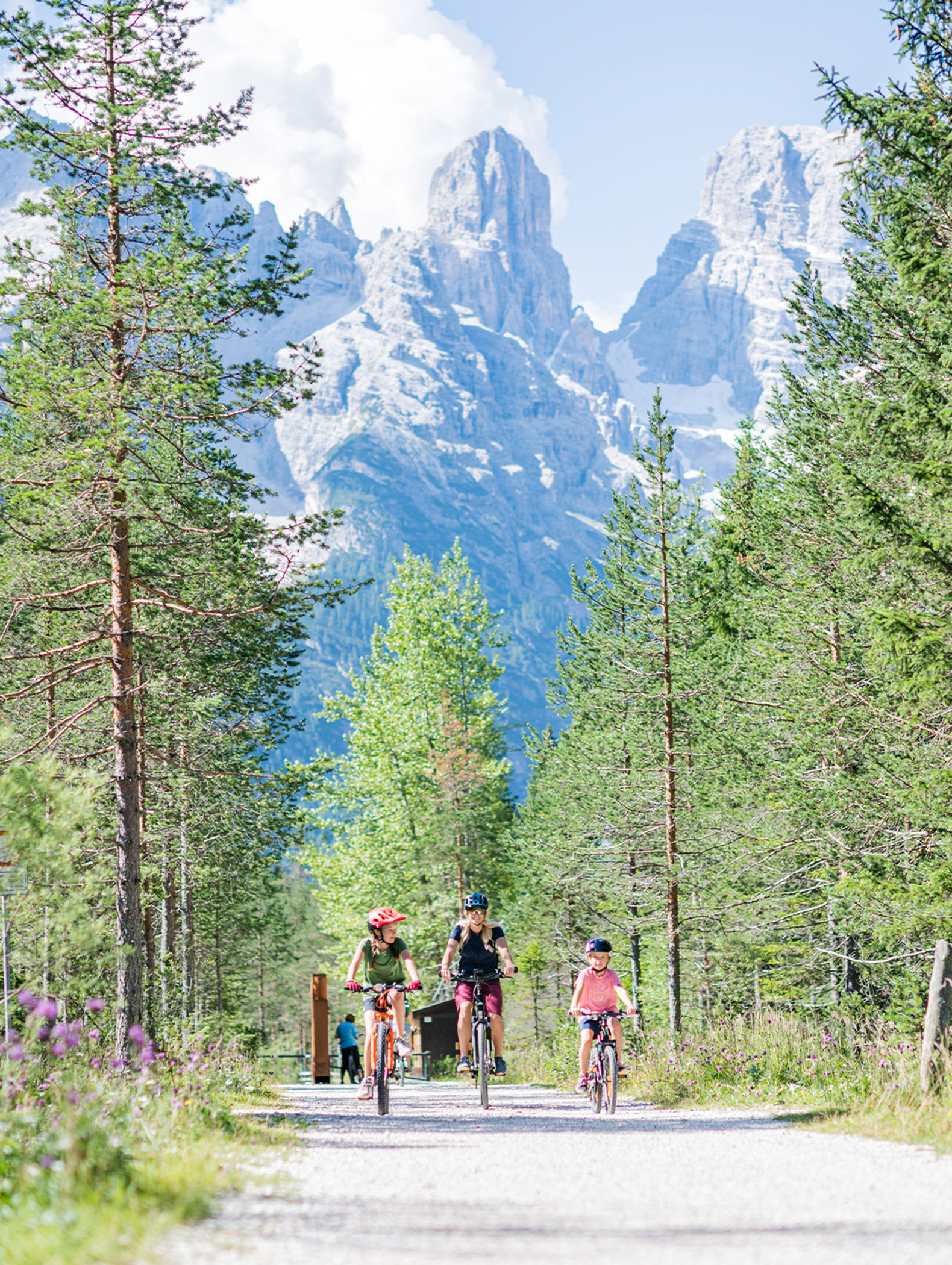 Drei Personen fahren an einem sonnigen Tag auf einem Waldweg mit hohen Bergen im Hintergrund Fahrrad. - Majestic Resorts (Relaunch)