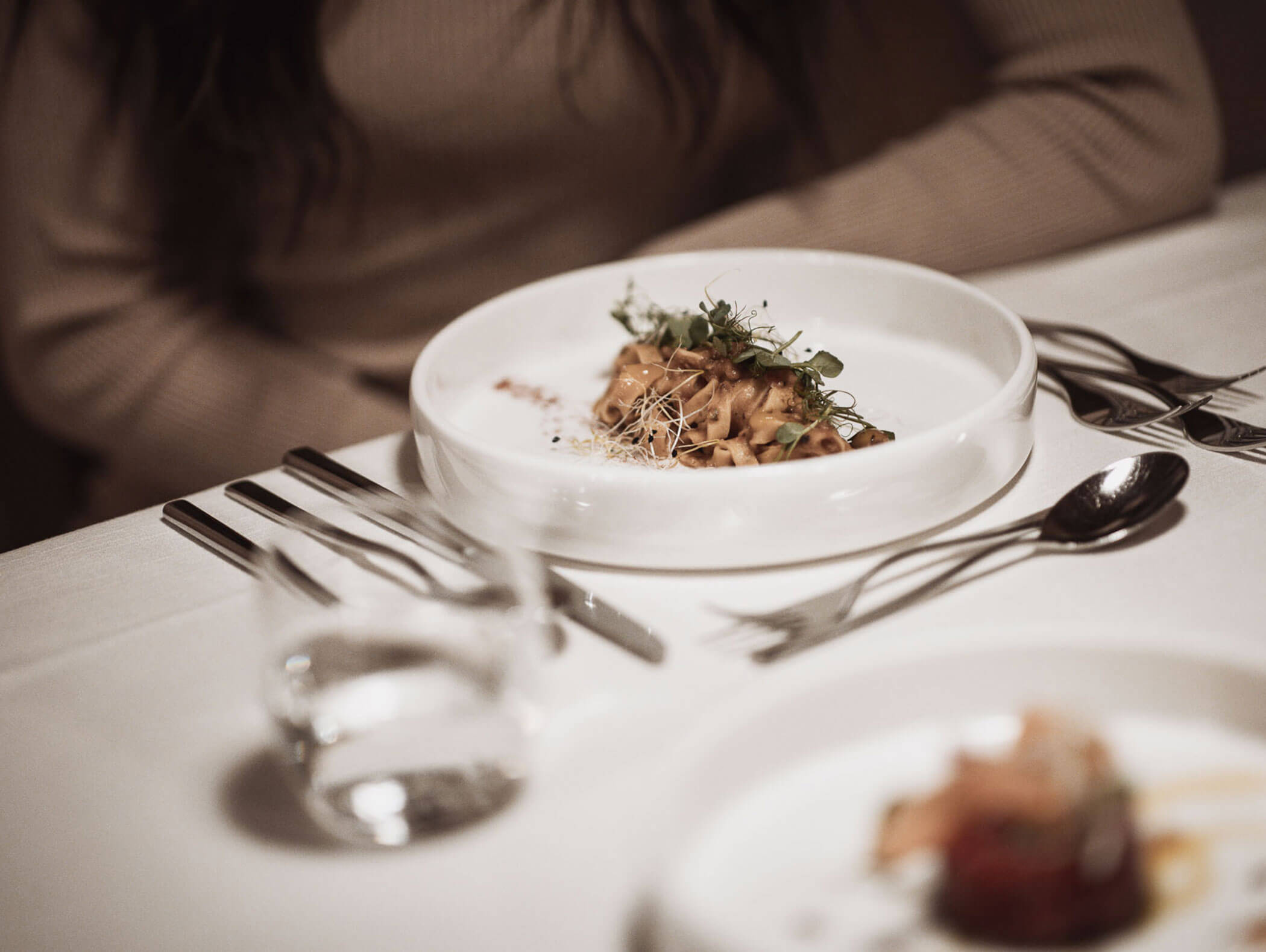 Eine Person sitzt an einem Tisch mit einem Teller mit Essen, Besteck und einem Glas Wasser auf einem weißen Tischtuch. - Majestic Resorts (Relaunch)
