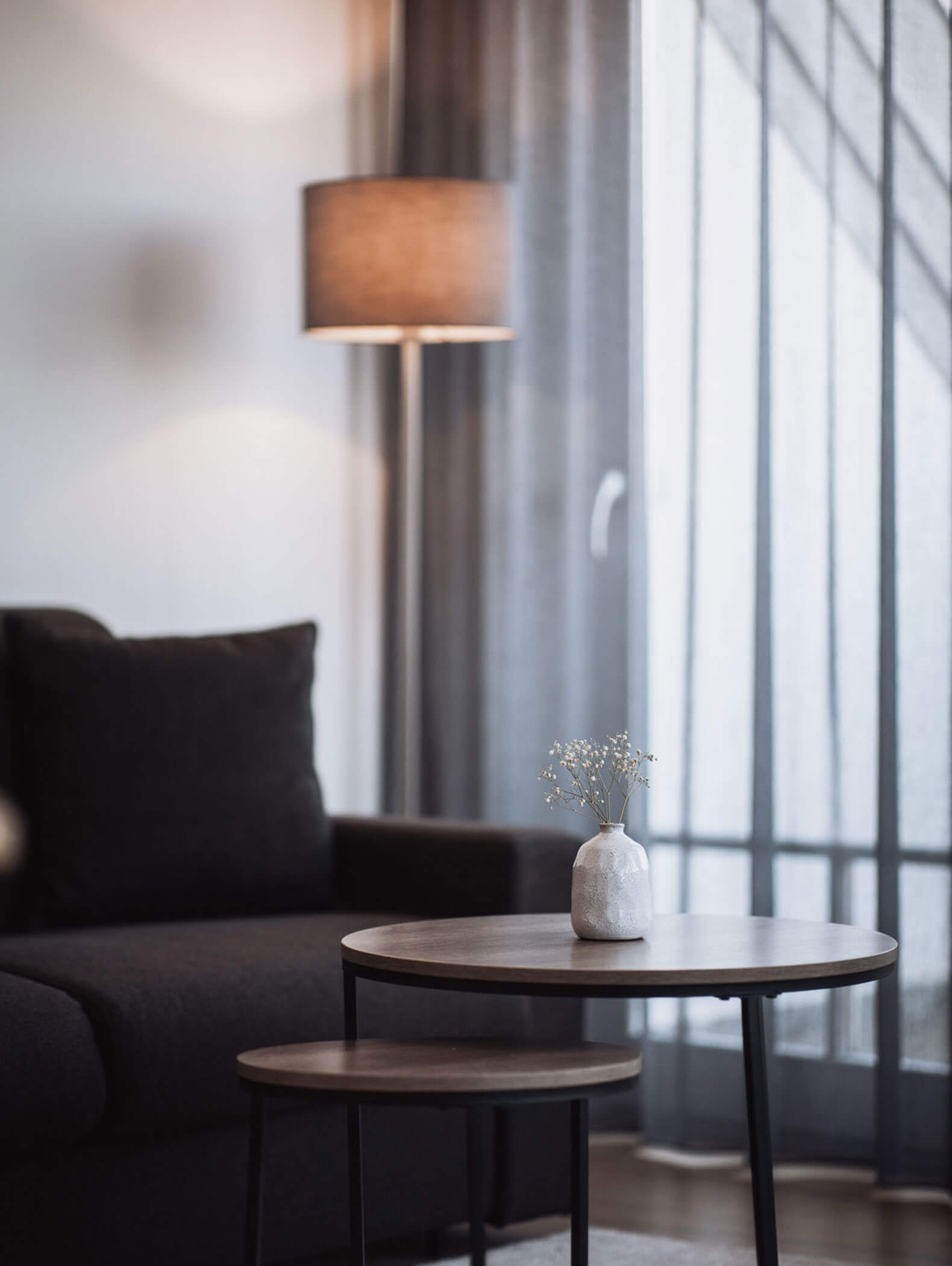 Minimalistisches Wohnzimmer mit runden Beistelltischen, einer kleinen weißen Vase, einem dunklen Sofa und durchsichtigen Vorhängen. - Majestic Resorts (Relaunch)