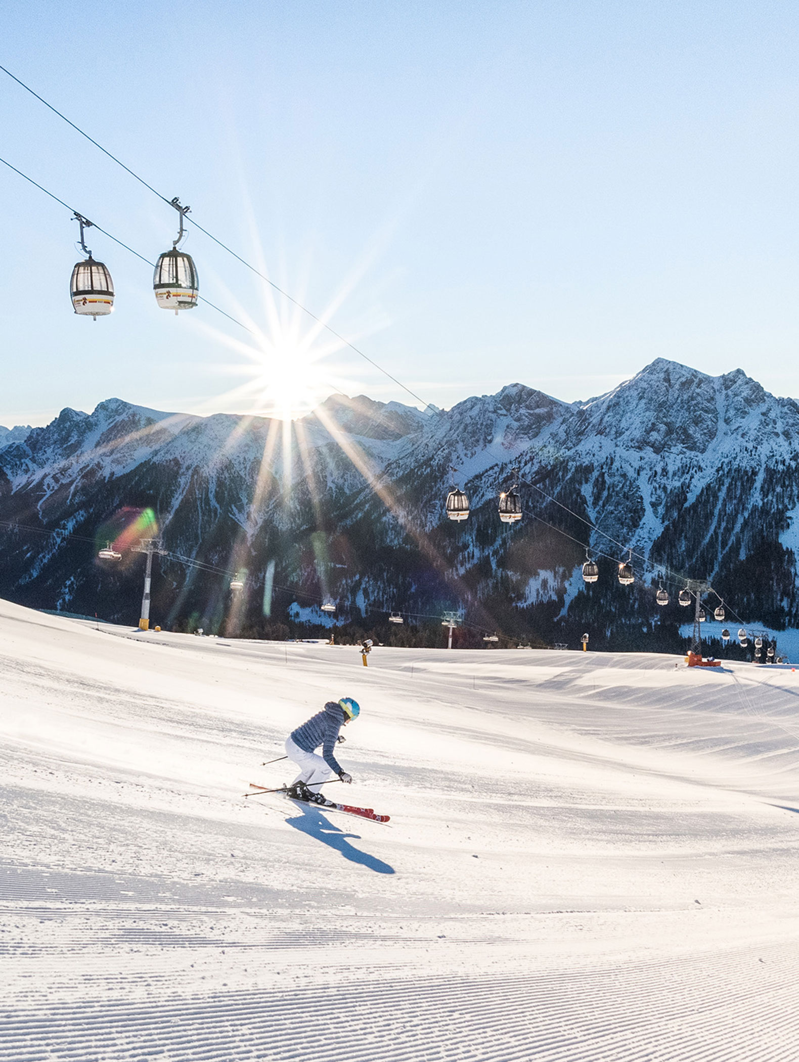 Skifahrer auf einer verschneiten Piste mit Skiliften über ihnen und der aufgehenden Sonne über den schneebedeckten Bergen im Hintergrund. - Majestic Resorts (Relaunch)