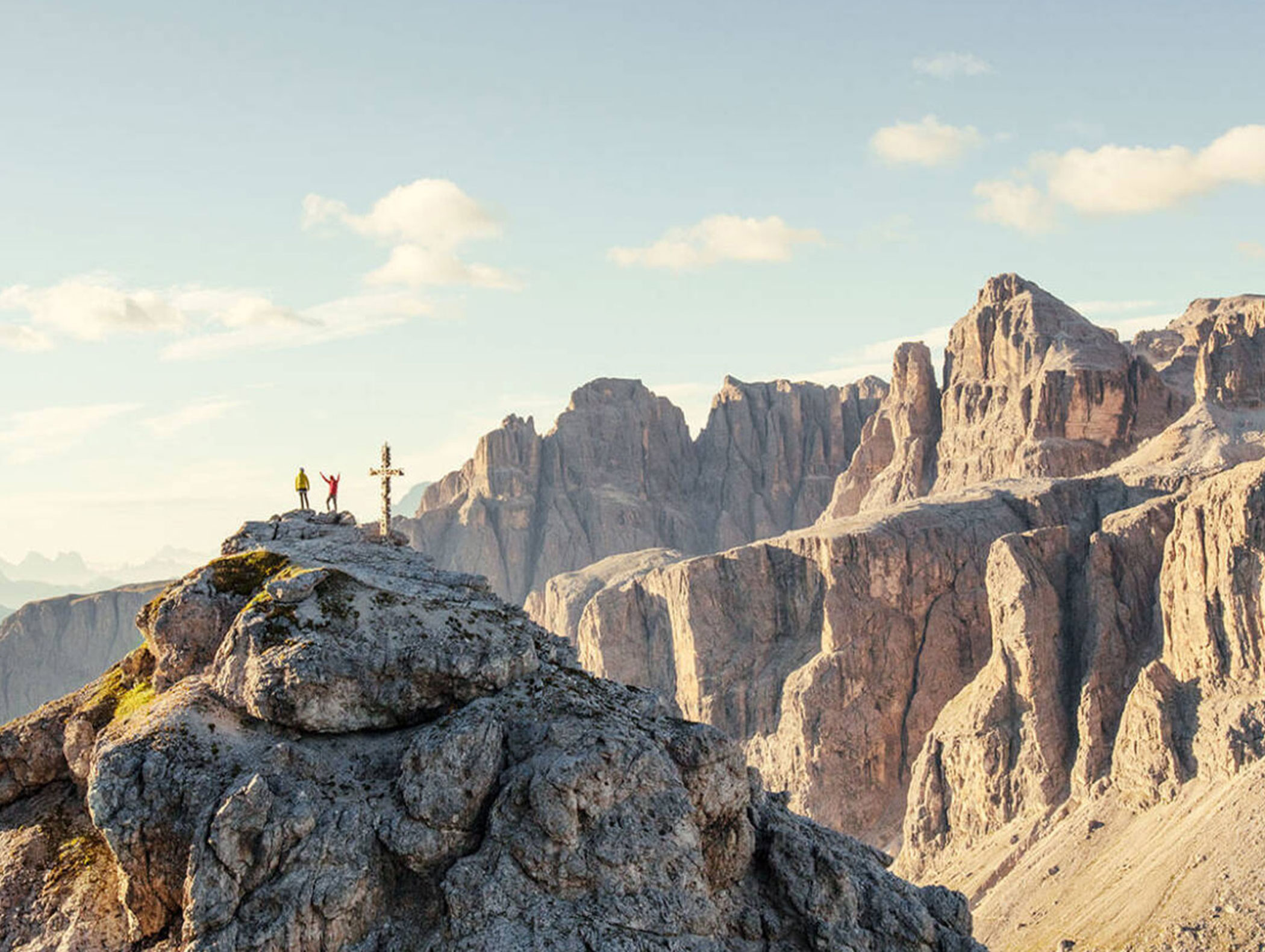 Drei Wanderer stehen auf einem felsigen Berggipfel in der Nähe eines Kreuzes, umgeben von dramatischen Felsen unter einem klaren Himmel. - Majestic Resorts (Relaunch)