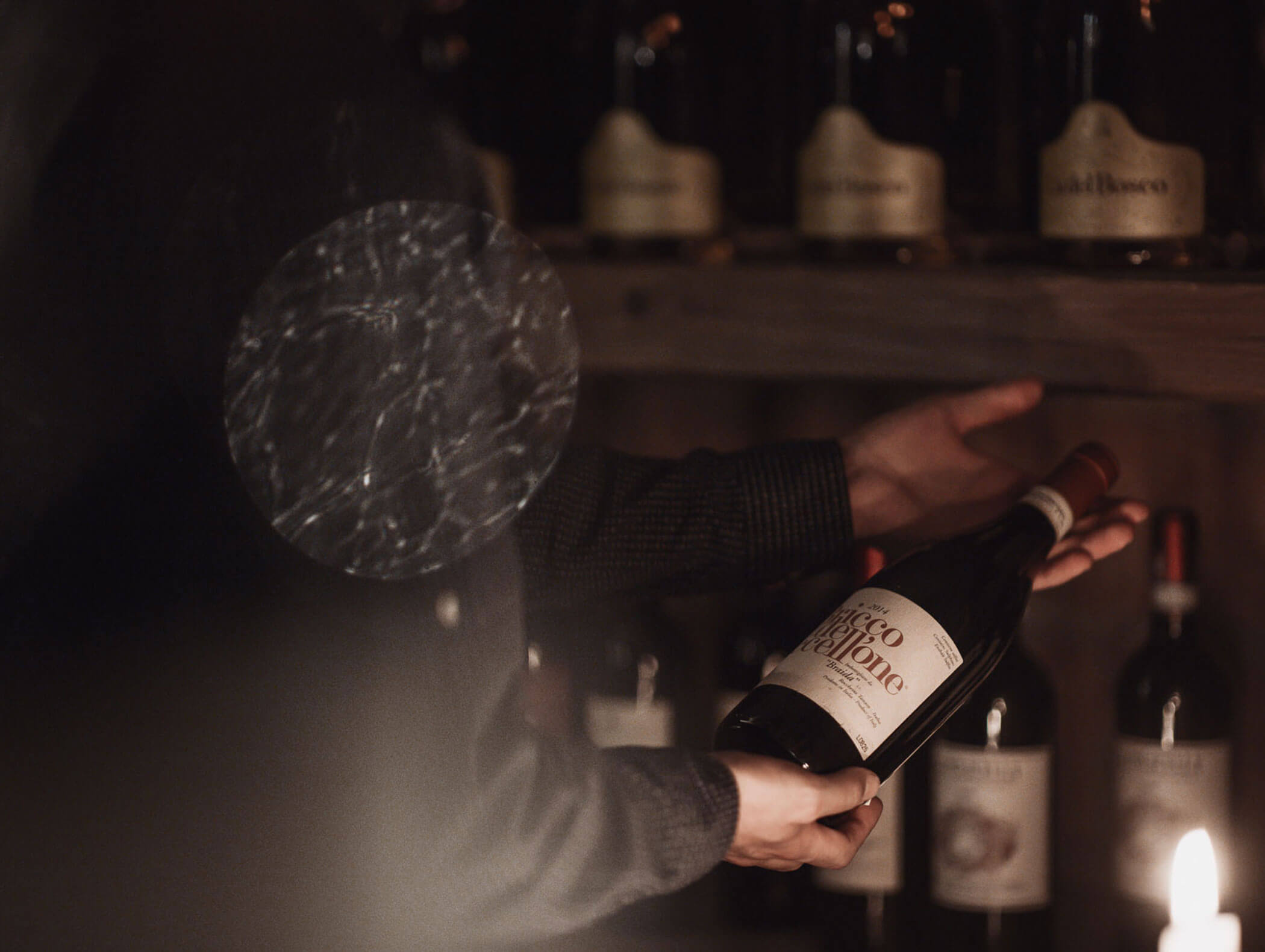 Eine Person hält eine Flasche Rotwein in einem schwach beleuchteten Weinkeller, mit Regalen voller Flaschen im Hintergrund. - Majestic Resorts (Relaunch)