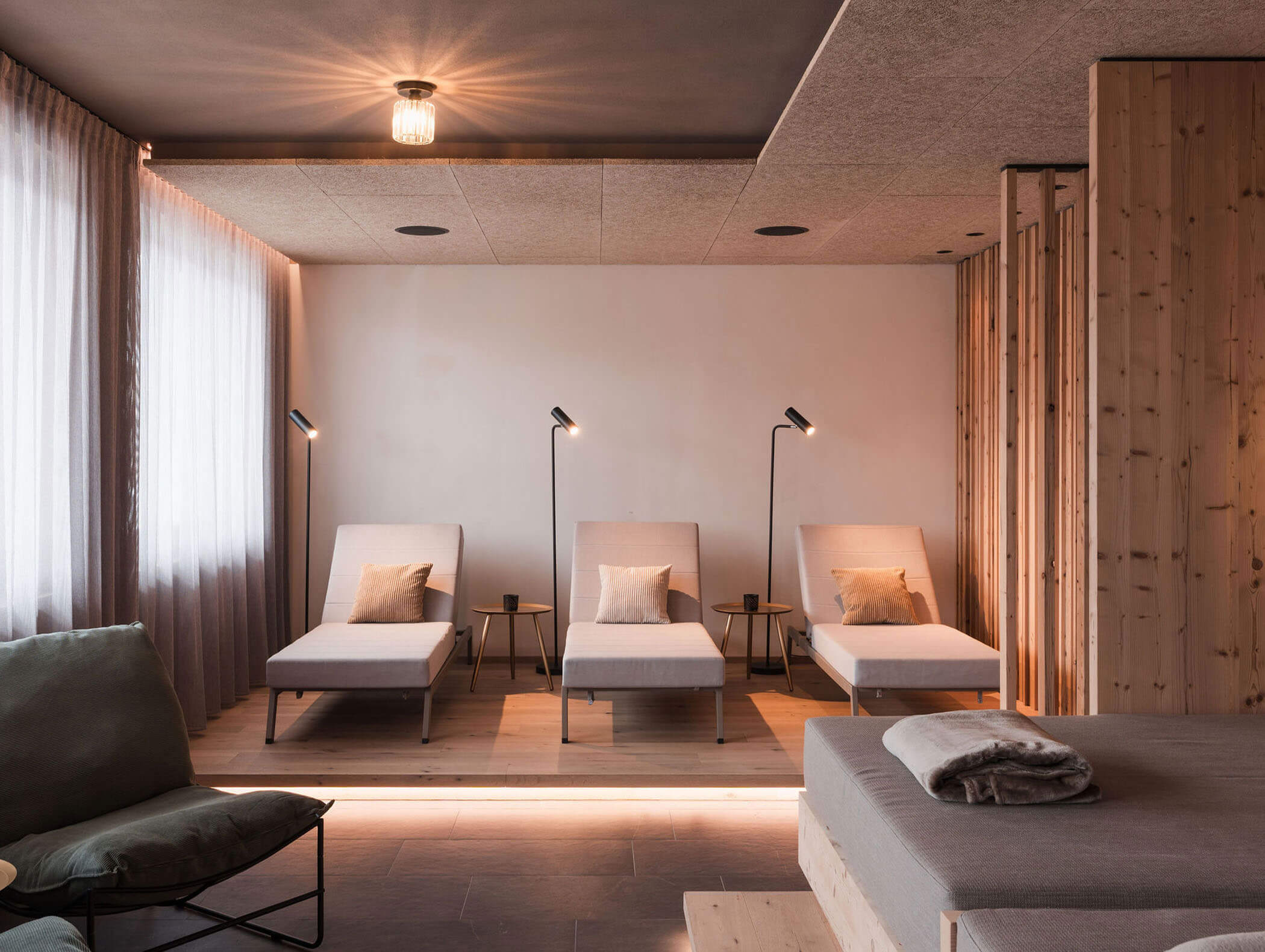 Drei moderne Lounge-Sessel mit Kissen und Beistelltischen in einem ruhigen, sanft beleuchteten Spa-Raum mit Holzakzenten. - Majestic Resorts (Relaunch)