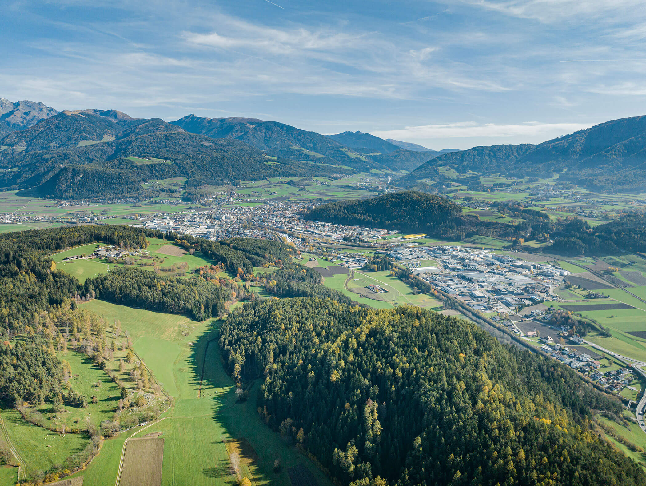 Luftaufnahme eines grünen Tals mit Wäldern, Feldern und einer von Bergen umgebenen Stadt unter blauem Himmel. - Majestic Resorts (Relaunch)