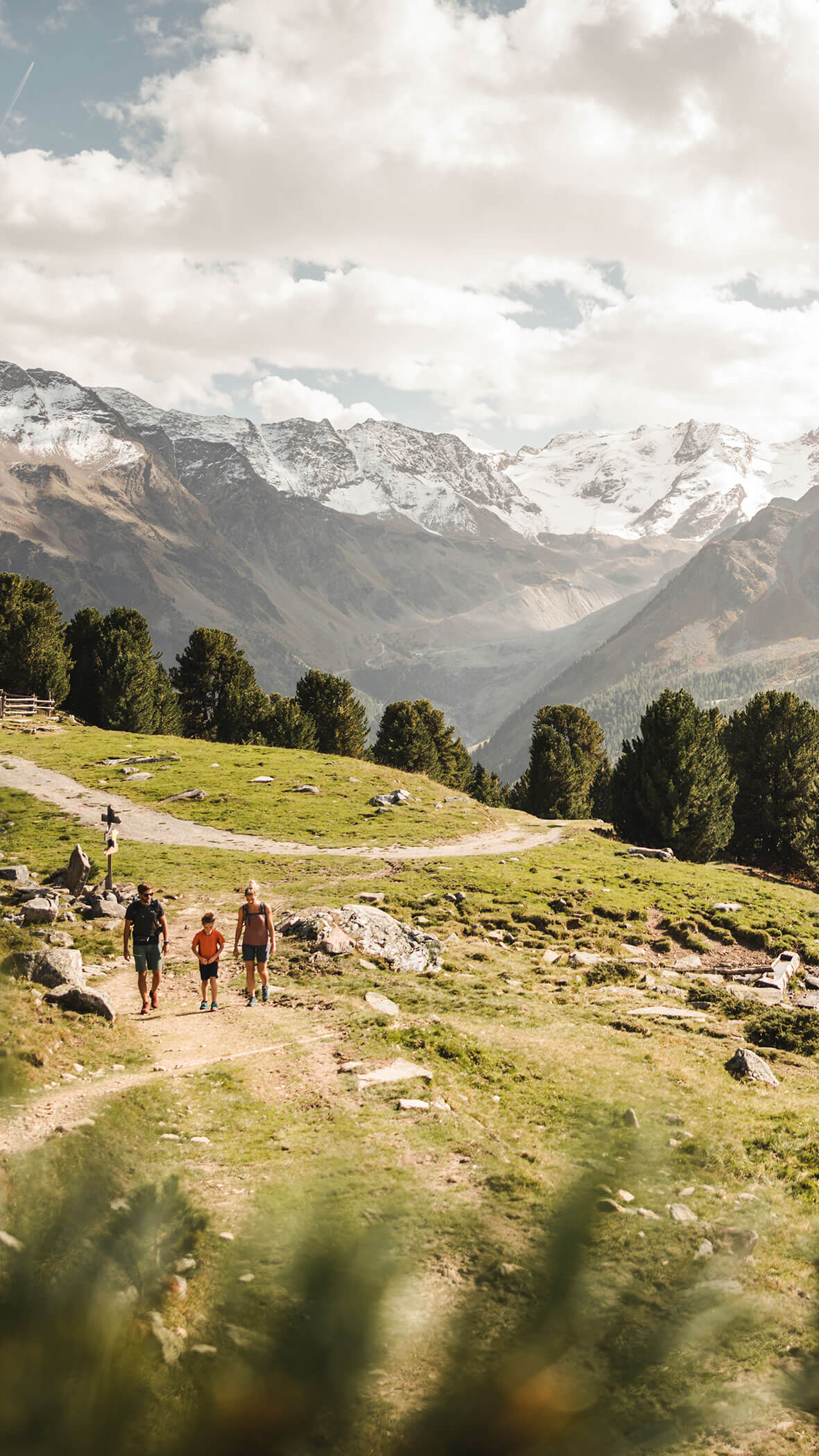 Vier Personen wandern auf einem Bergpfad mit schneebedeckten Gipfeln und grünen Bäumen im Hintergrund. - Majestic Resorts (Relaunch)