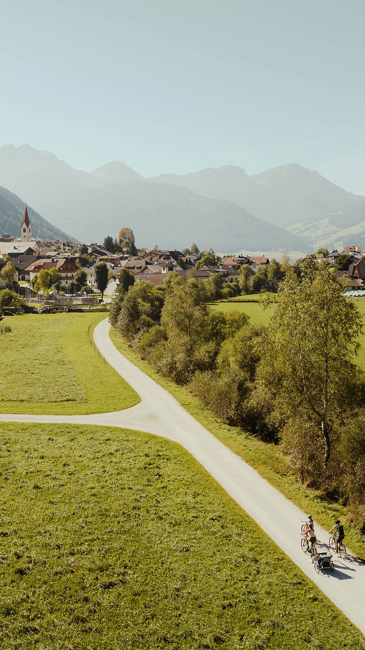 Ein gewundener Weg führt zu einem Dorf mit Bergen im Hintergrund; drei Personen fahren mit dem Fahrrad auf dem Weg. - Majestic Resorts (Relaunch)