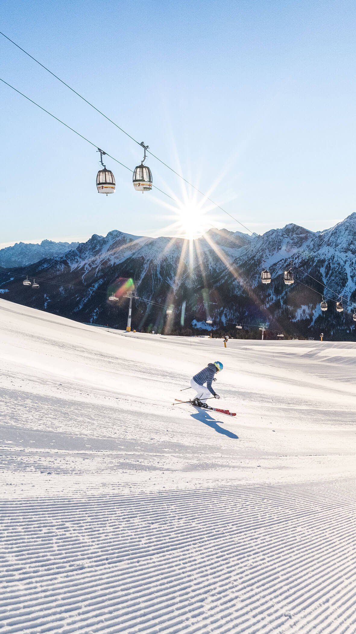 Skifahrer auf einer präparierten Schneepiste mit strahlender Sonne, Bergen und Seilbahnen im Hintergrund. - Majestic Resorts (Relaunch)