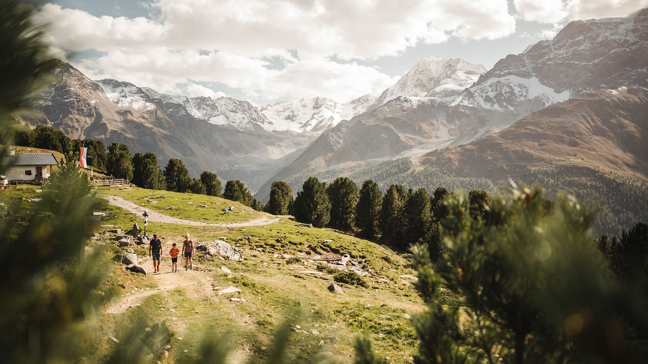 Eine Gruppe von Menschen wandert auf einem Bergpfad mit verschneiten Gipfeln und grünen Bäumen im Hintergrund. - Majestic Resorts (Relaunch)