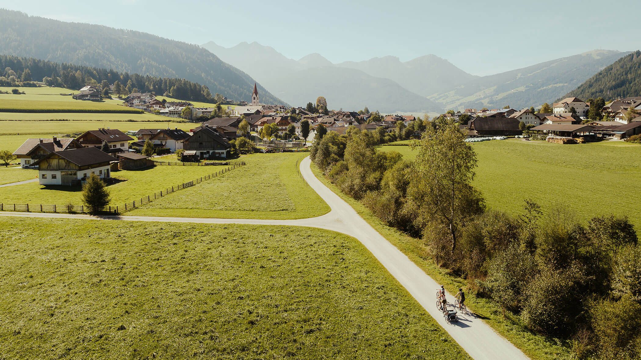 Eine kurvenreiche Straße durch grüne Felder führt zu einem Dorf mit Bergen im Hintergrund; zwei Radfahrer fahren auf der Straße. - Majestic Resorts (Relaunch)