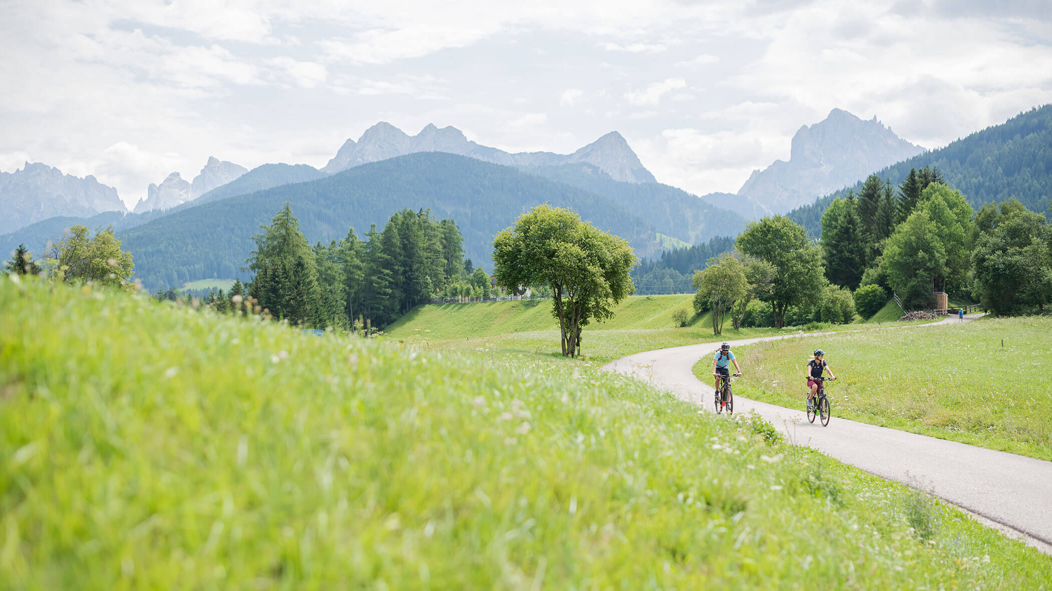 Zwei Radfahrer fahren auf einem gewundenen Weg durch grüne Wiesen, mit Bergen und Bäumen im Hintergrund. - Majestic Resorts (Relaunch)