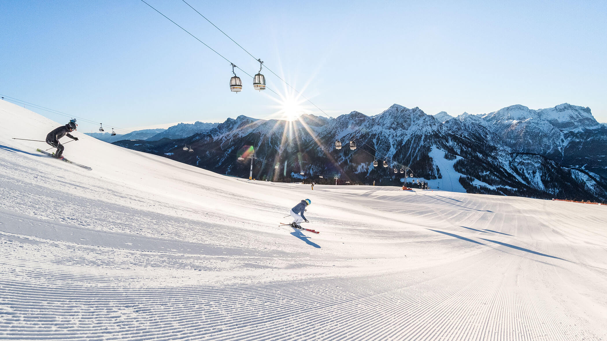 Zwei Skifahrer fahren eine sonnige, präparierte Piste hinunter, mit Bergen und Skiliften im Hintergrund. - Majestic Resorts (Relaunch)