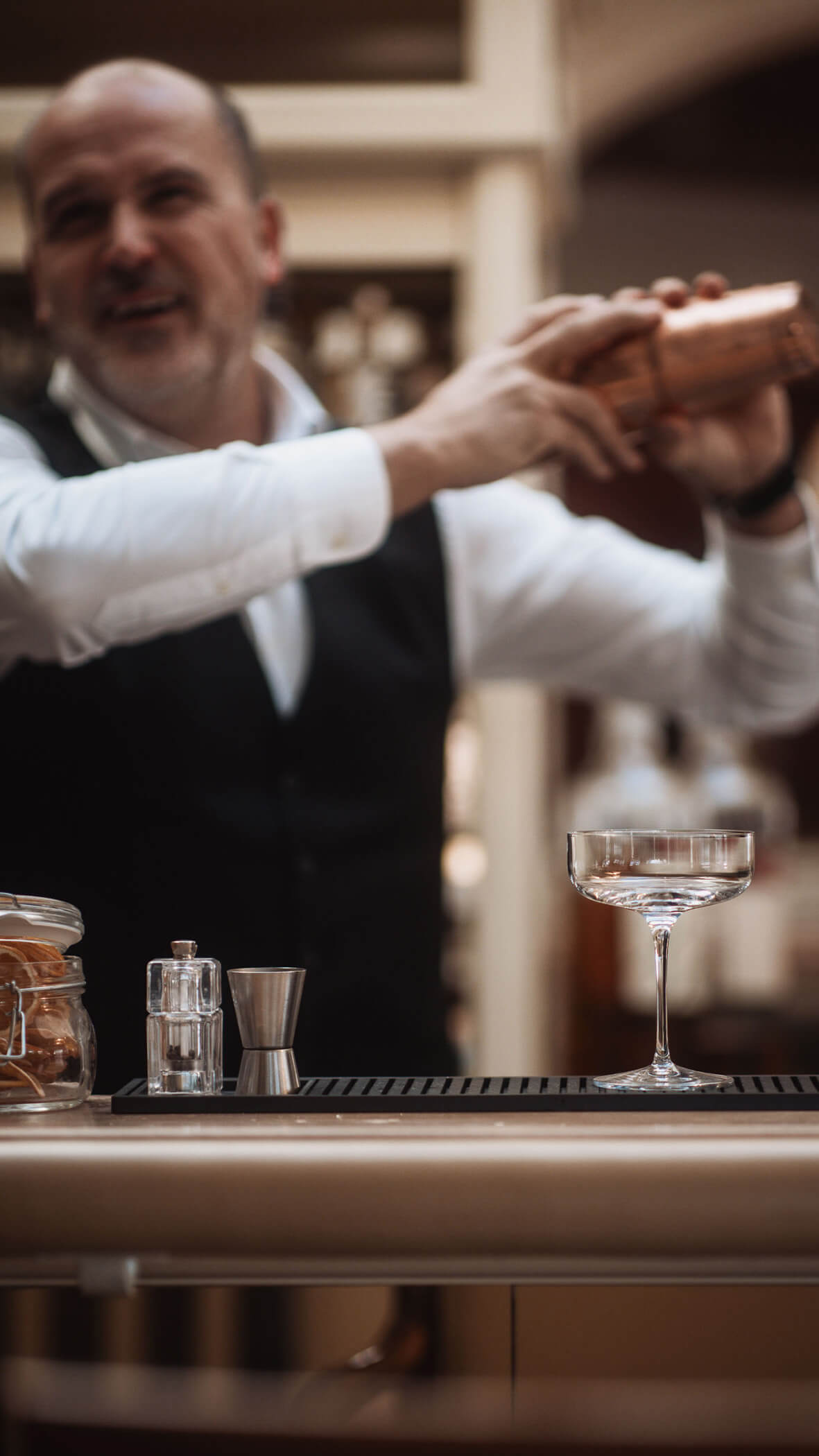 Barkeeper in Weste schüttelt einen Cocktail-Shaker hinter einer Bar mit einem Glas und Gläsern im Vordergrund. - Majestic Resorts (Relaunch)
