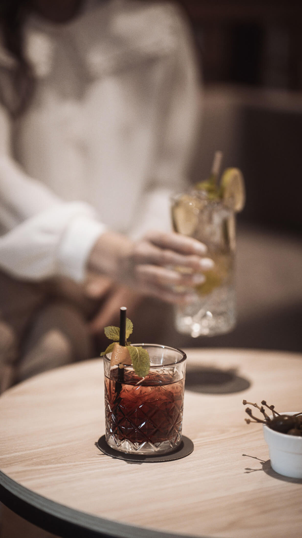 Eine Person greift nach einem Cocktailglas, auf dem Tisch steht ein anderes Getränk, garniert mit Minze und Orange. - Majestic Resorts (Relaunch)