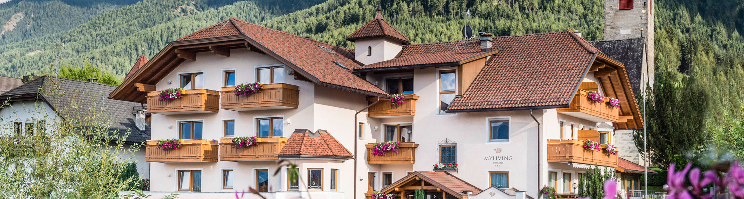 Hotel im alpinen Stil mit Holzbalkonen und Blumenkästen inmitten einer bewaldeten Bergkulisse. - Majestic Resorts (Relaunch)